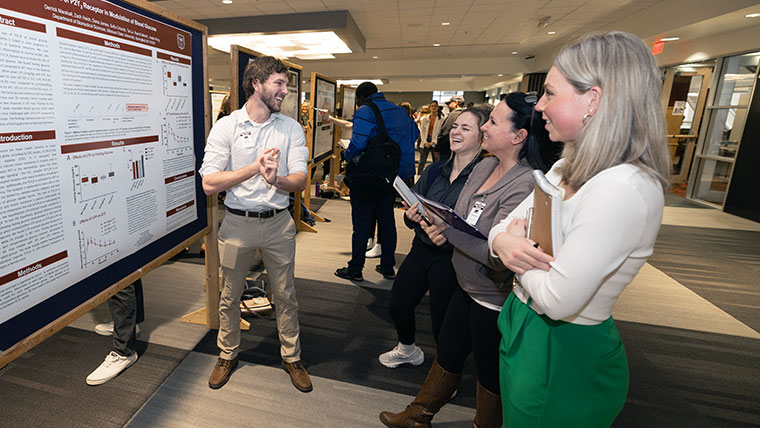 A smiling graduate student discussing his poster presentation with three individuals. 