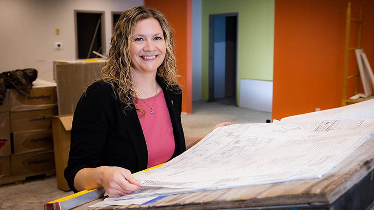 Smiling individual standing at a desk with architectural drawings on it. 